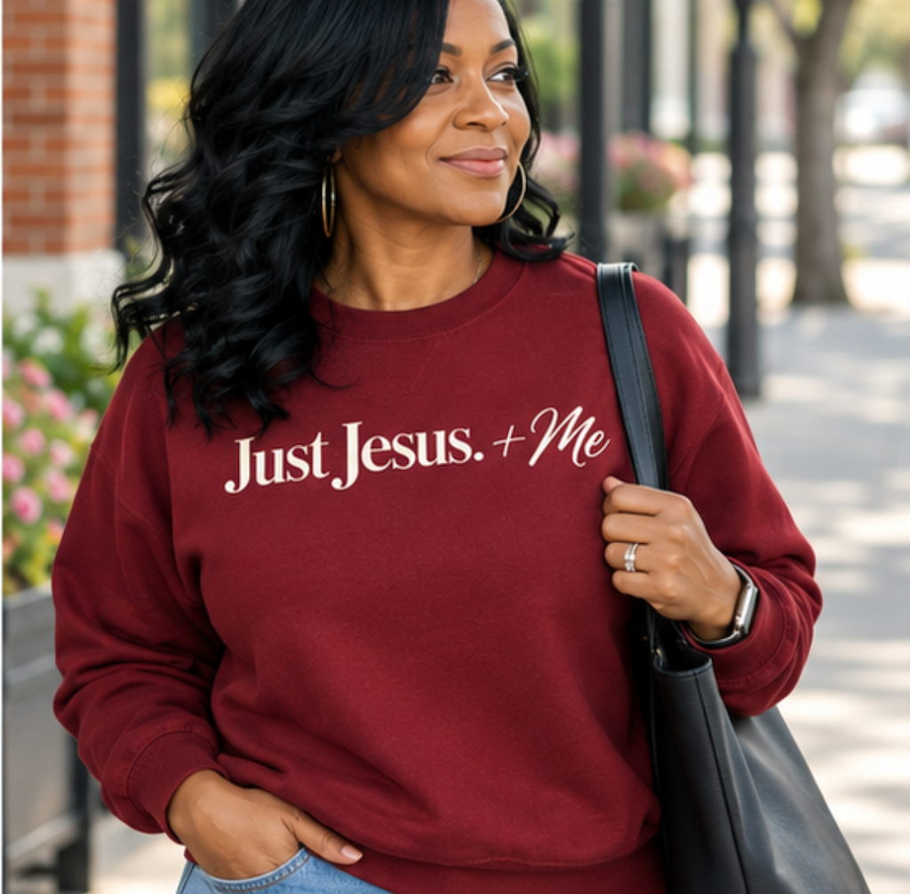 Black woman wearing the Just Jesus + Me garnet crewneck sweatshirt with cream lettering and light-wash jeans, enjoying a casual afternoon on a neighborhood sidewalk with brick storefronts and greenery in the background.