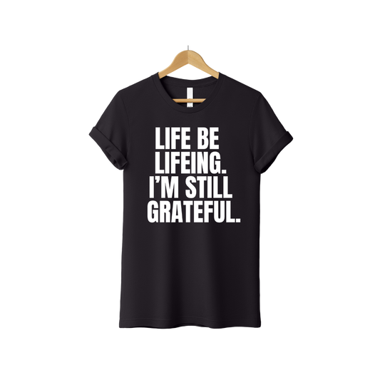 Black Christian T-shirt with bold white text reading “Life Be Lifeing. I’m Still Grateful.” faith-based encouragement tee and gratitude message.